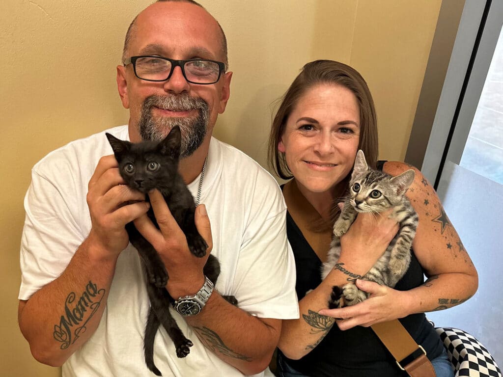A man with glasses and tattoos smiles while holding a black kitten in his left arm. Beside him, a woman with light brown hair holds a gray tabby kitten. They are both in a well-lit room with a neutral-colored wall.