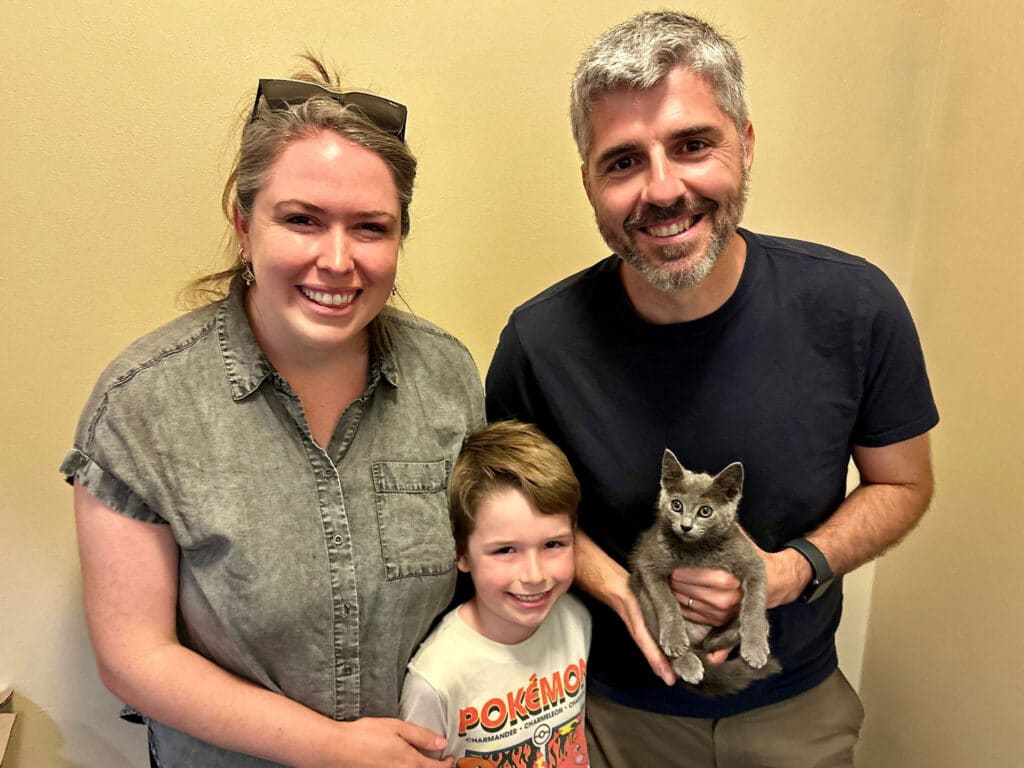 A woman in a gray shirt, a boy in a white shirt with a Pokémon graphic, and a man in a black shirt stand together. The man holds a small gray kitten. They are in a room with a light-colored wall.