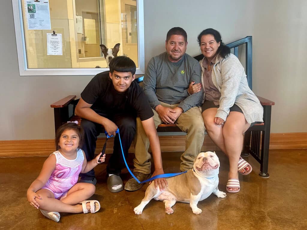 A family of four sits on a bench in an adoption center. A young girl in a pink outfit sits in front, while a teenage boy with a leash stands beside a seated man holding a dog. A woman sits next to the man, smiling. A dog's head is visible on the floor.