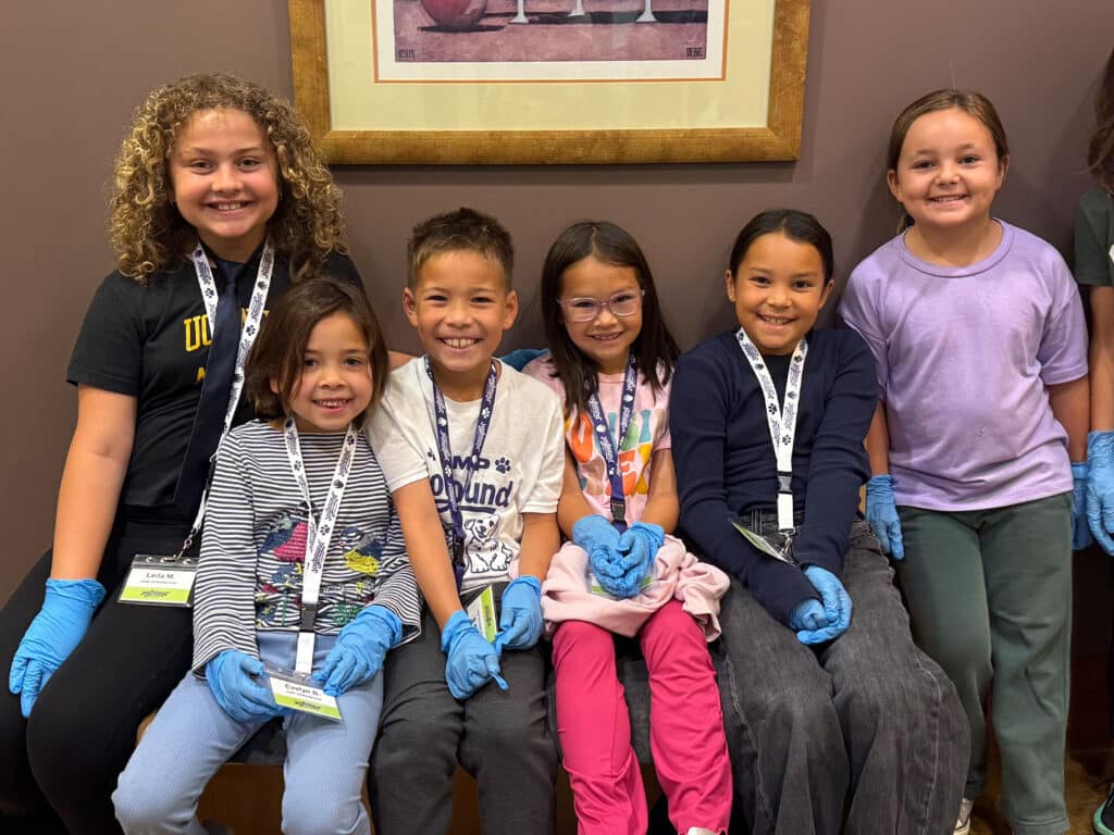 A group of six children wearing gloves sit together on a bench. They are dressed in various colorful shirts and pants, and some have name tags. A framed picture is visible on the wall behind them.