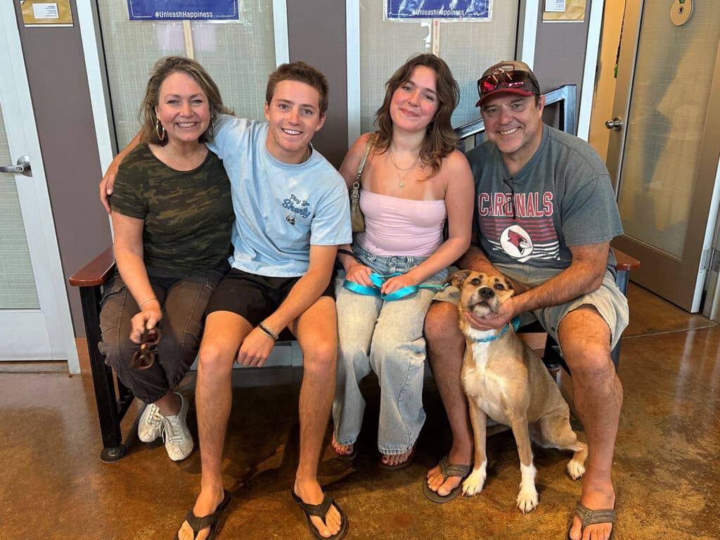 A family of four and a tan dog are seated together on a bench in a room. The woman on the left wears a black shirt, and the man on the right wears a gray t-shirt. Two teenagers sit between them, one in a light blue shirt and the other in a pink top.