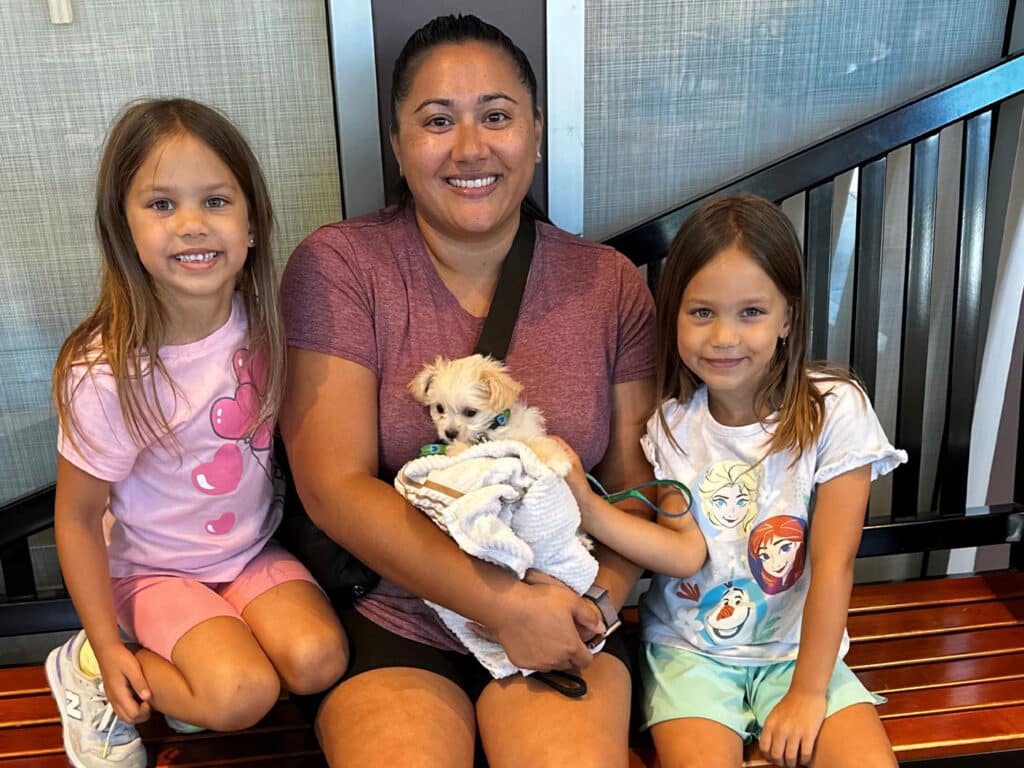 A woman holding a small puppy wrapped in a white blanket sits on a bench. Beside her are two girls, one in a pink shirt and shorts and the other in a white shirt with characters on it, both smiling. The background features a modern interior.