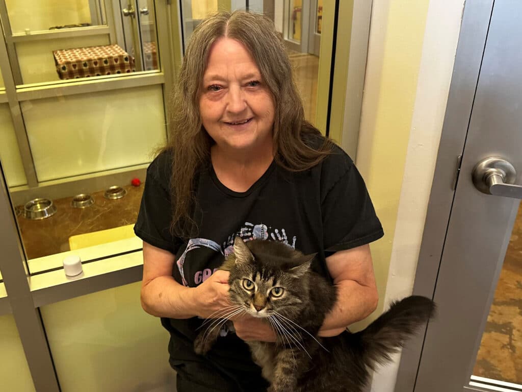 A woman with long brown hair, wearing a black T-shirt, sits on the floor holding a gray and black striped cat. In the background, several empty food bowls are visible inside a glass enclosure.