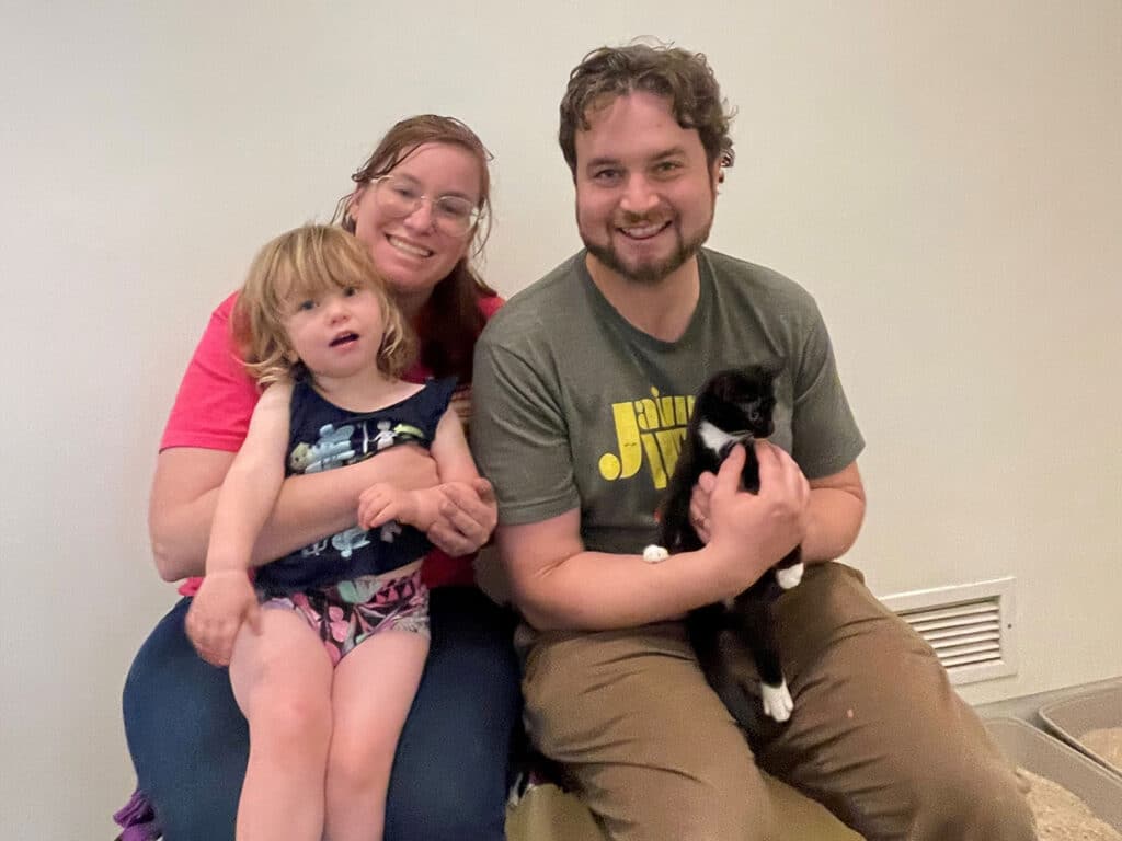 A woman with long brown hair and glasses sits on a stool holding a young girl in a colorful swimsuit. Beside them, a man with curly hair and a gray shirt holds a black and white cat. They are seated against a plain wall.