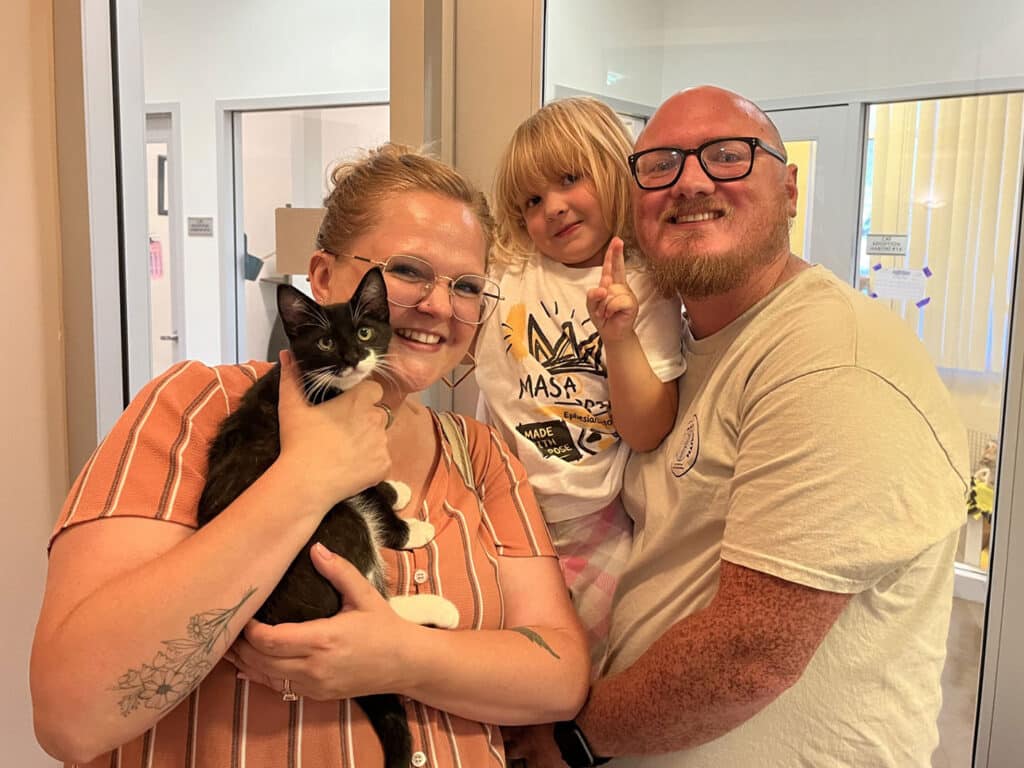 A woman holding a black and white kitten stands beside a man with glasses. A child is seated in the man's arms, making a peace sign. The background features large windows with natural light.