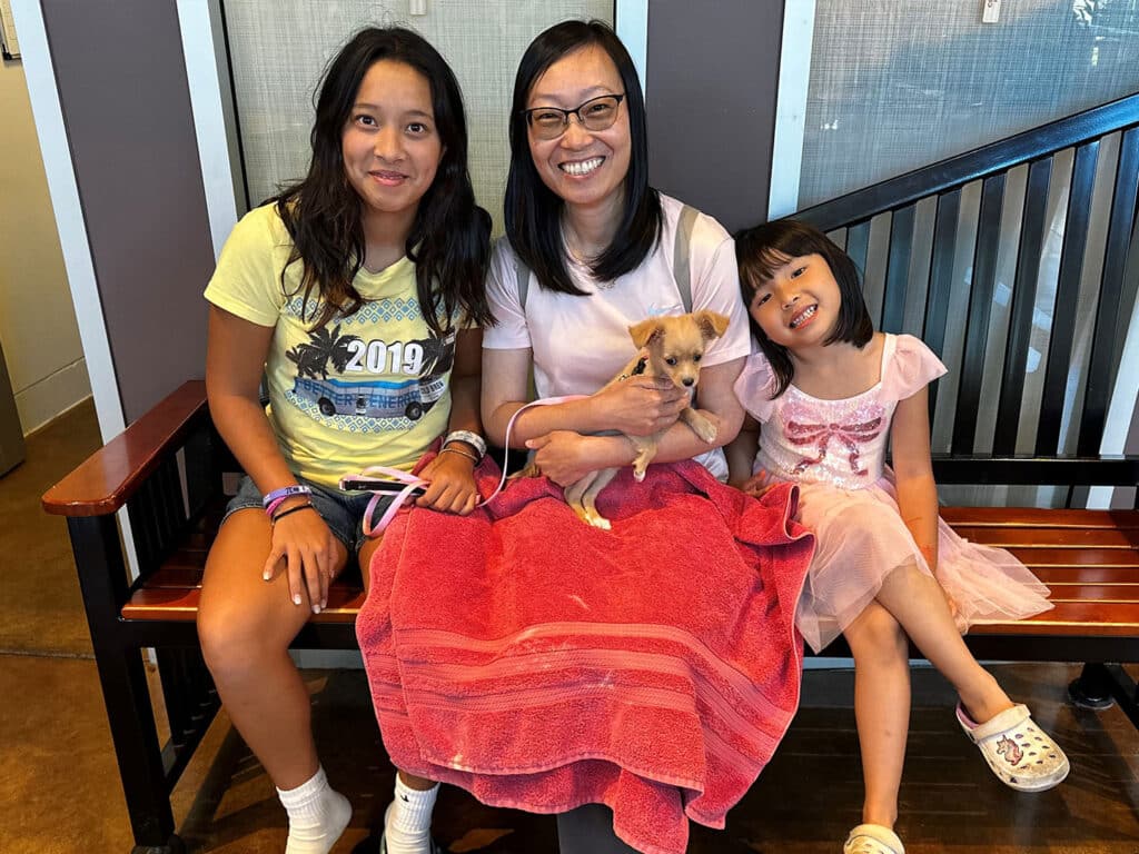 A woman wearing glasses and a pink shirt sits on a bench, holding a small tan dog. Beside her are two girls, one in a light green shirt and the other in a pink dress, both smiling at the camera. The background features wooden slats.