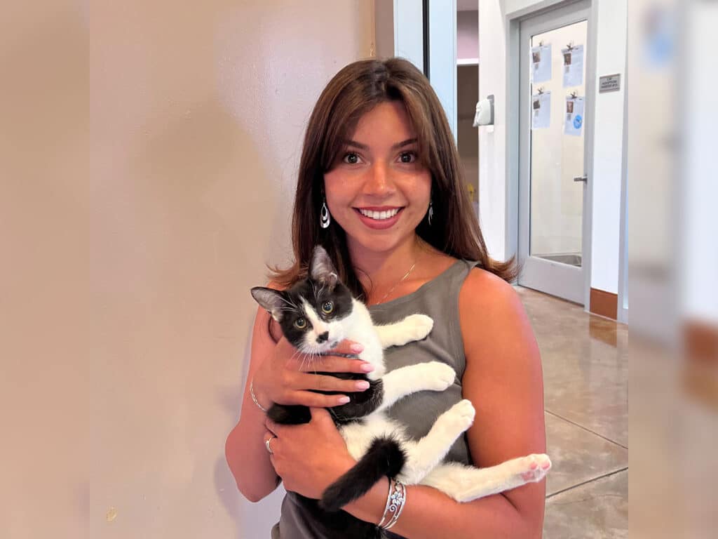 A woman with long brown hair is holding a black and white kitten in her arms. She is wearing a gray sleeveless top and earrings. The background features light-colored walls and a door leading to another room.