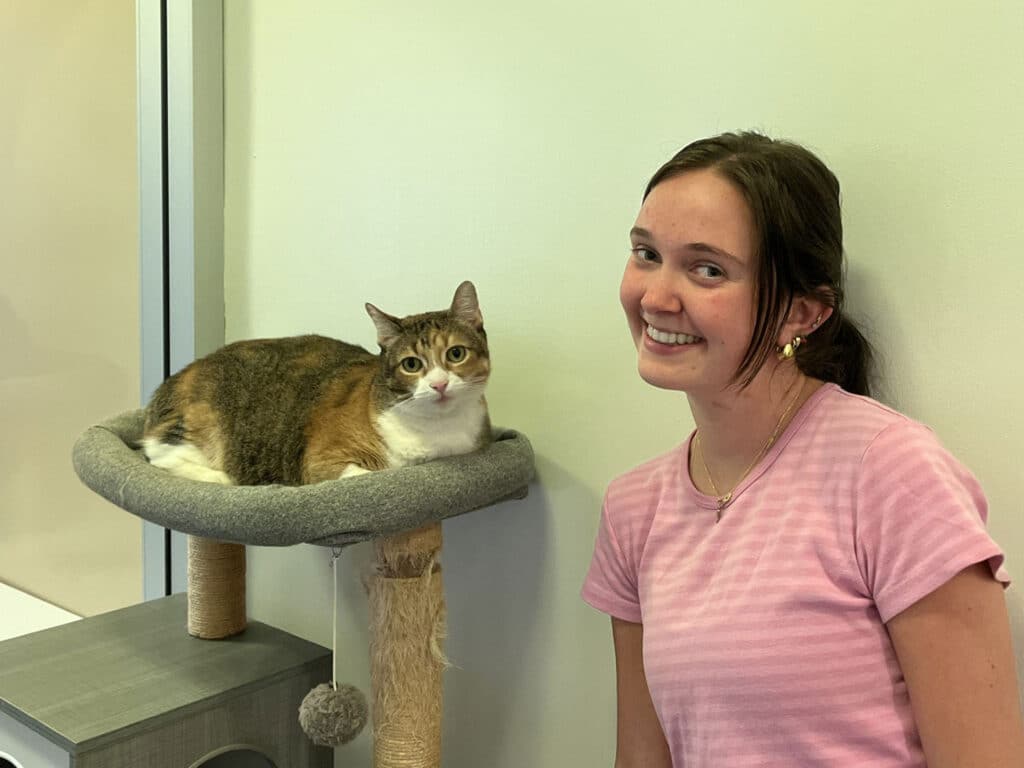A young woman with long dark hair and wearing a pink shirt smiles while sitting next to a cat on a gray perch. The cat, with a mix of brown and white fur, rests comfortably on the perch against a light-colored wall.