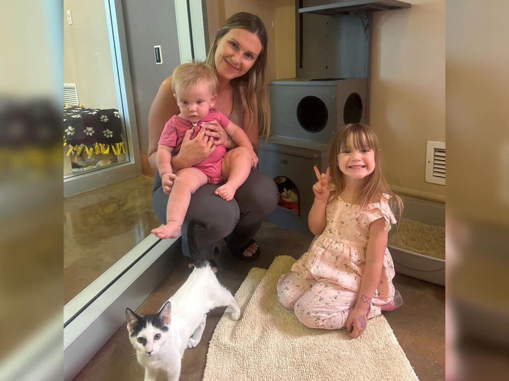 A woman holding a toddler sits on the floor beside a girl with long hair, who is holding up two fingers in a peace sign. A small black and white kitten is sitting on a beige rug in front of them. The background includes glass walls and an animal care area.