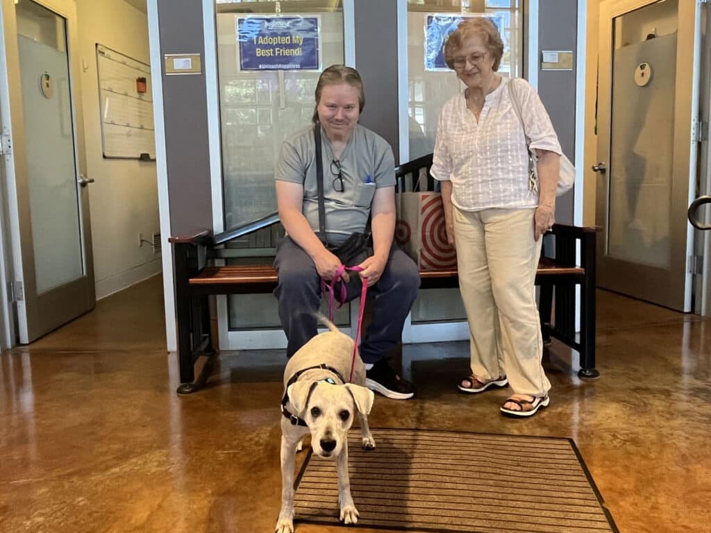 A man seated on a bench holds a leash connected to a light-colored dog, while a woman stands beside him smiling. The background features large windows, and a sign that reads "Adoption: Best Friend" is visible on the wall.