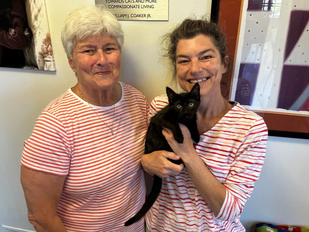 Two women wearing red and white striped shirts stand together in an indoor setting. One woman, on the left, has short gray hair, while the other has long brown hair and is holding a small black kitten.