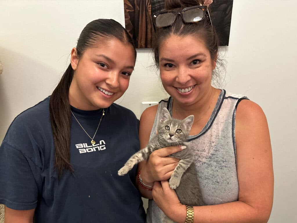A young woman with long hair and a navy t-shirt stands next to a smiling woman wearing a sleeveless top, holding a gray kitten. They are positioned in front of a wall with artwork and a white outlet.