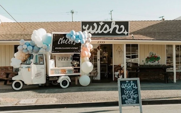 A small white food truck adorned with colorful balloons is parked in front of a shop with a sign reading "wish." A chalkboard sign displays promotions outside, while the shop features large windows and decorative plants.