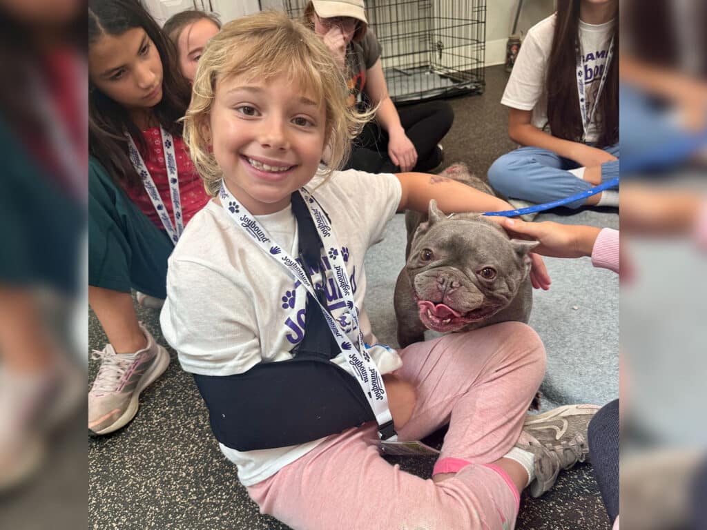 A young girl with short blonde hair smiles while sitting on the floor, wearing a white camp t-shirt and a black arm sling. She sits beside a gray dog with a flat face, and several other children are visible in the background.