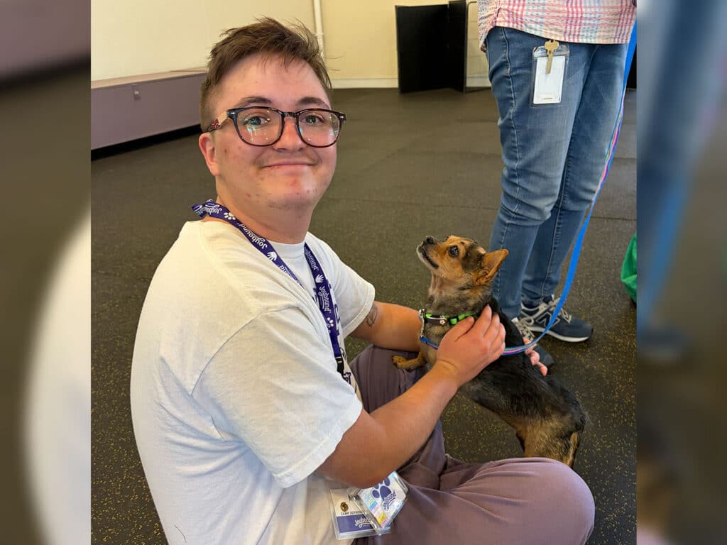 A person with glasses and short hair sits on the floor, gently holding a small dog in a harness. The dog looks up at the person. In the background, another person stands with their back to the camera.