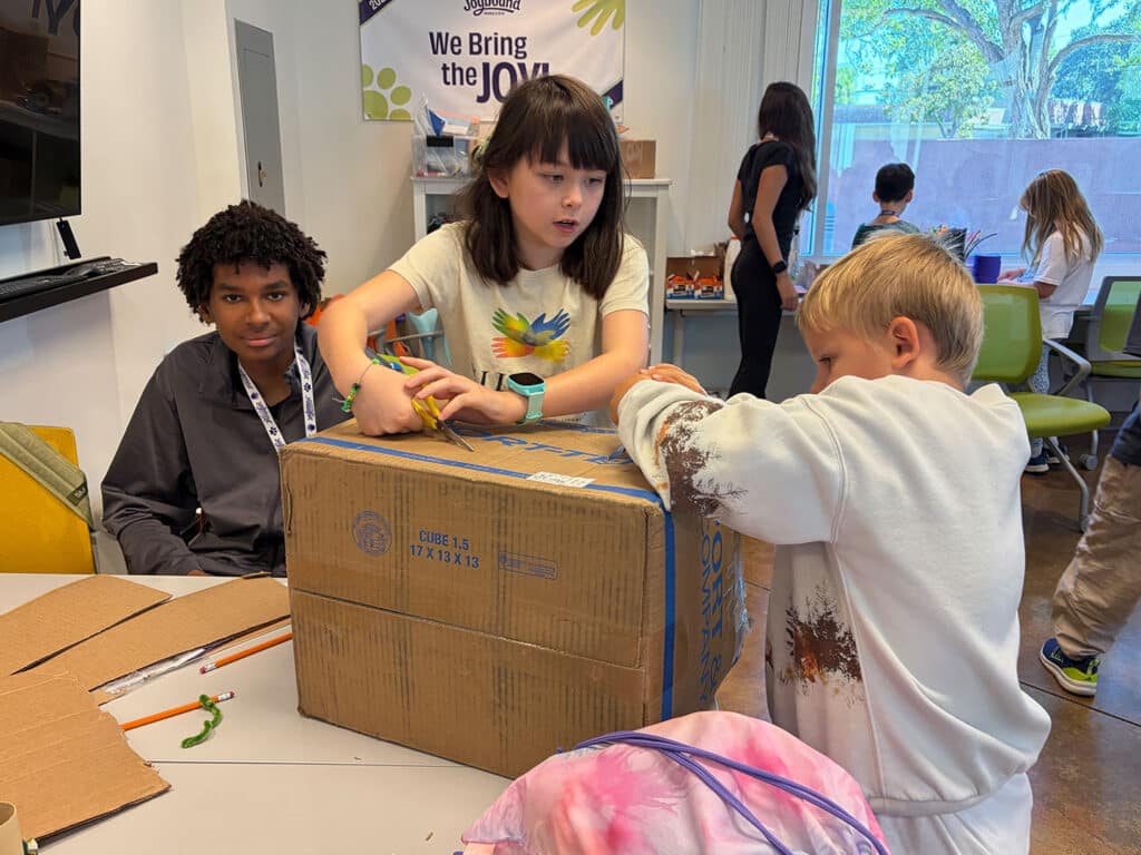 Three children work together to open a large cardboard box at a camp table, with markers, scissors, and a Camp Joybound banner visible in the room.