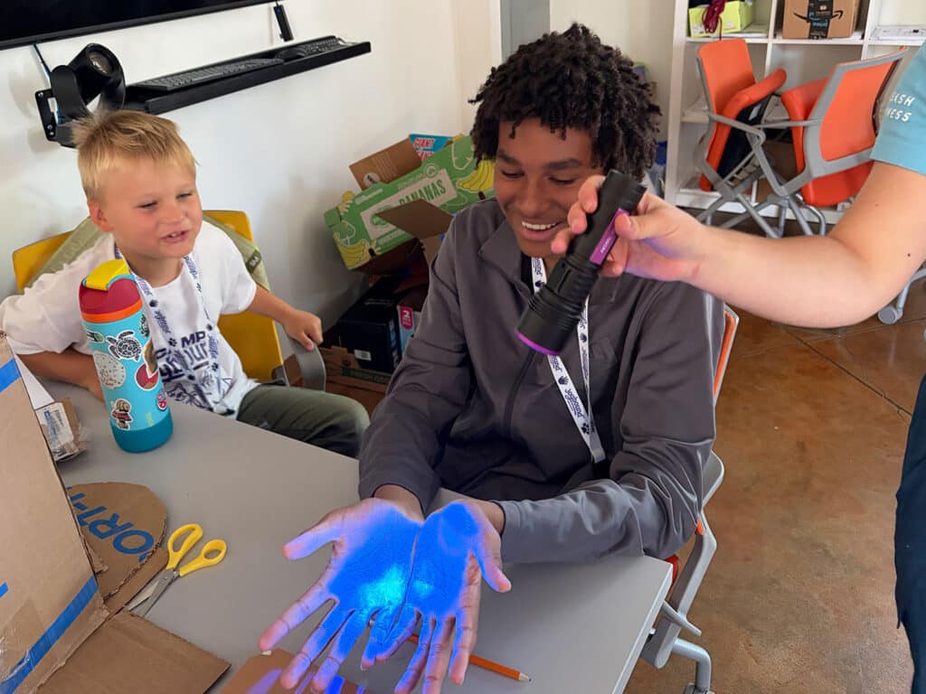A young man sits at a table with glowing blue light on his hands while another person shines a black flashlight on them, with a child and classroom supplies in the background.