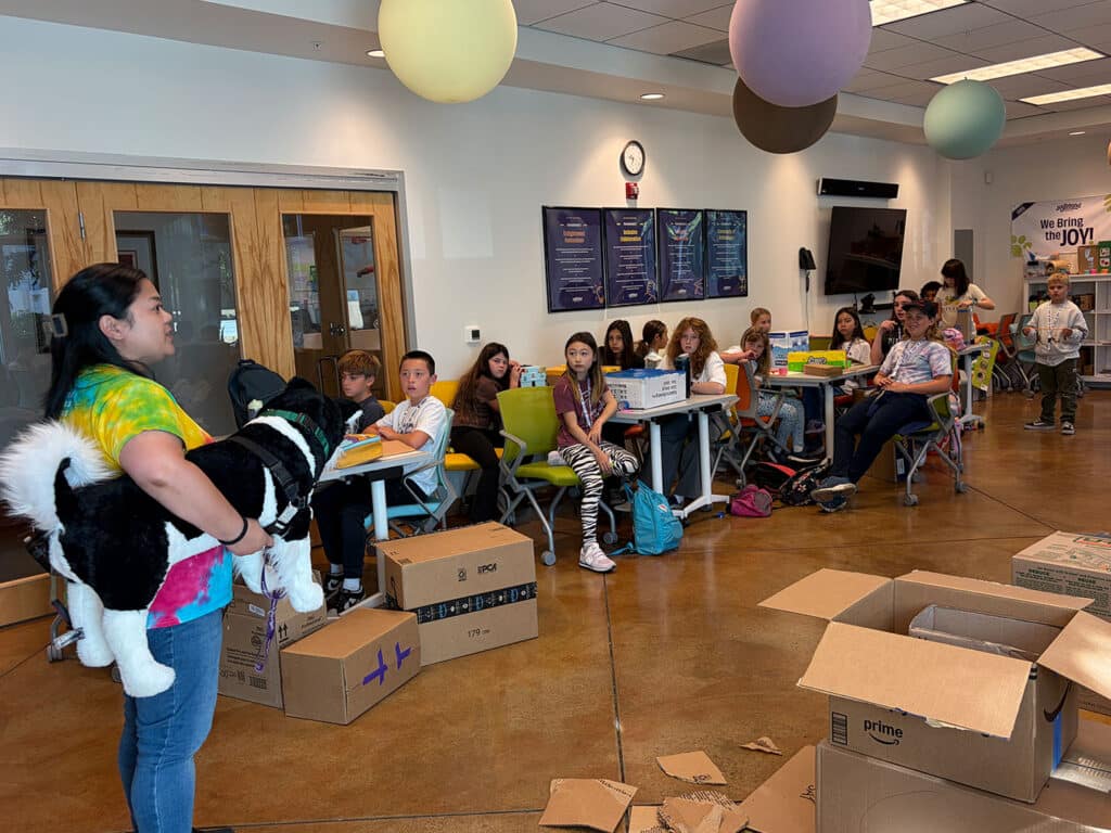 A woman in a tie-dye shirt holds a large black-and-white stuffed animal while seated children and counselors sit at tables in a camp room with hanging paper lanterns and cardboard boxes on the floor.