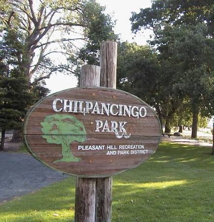 A wooden sign for Chilpancingo Park, which features the park's name in large white letters and a green tree icon. Below, it reads 'Pleasant Hill Recreation and Park District.' The sign is positioned amid trees and grassy areas.