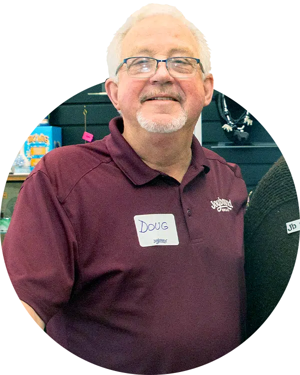 A man wearing glasses and a maroon polo shirt stands in front of a display with various items. He has a name tag that reads "Doug". The background consists of shelves with colorful merchandise.
