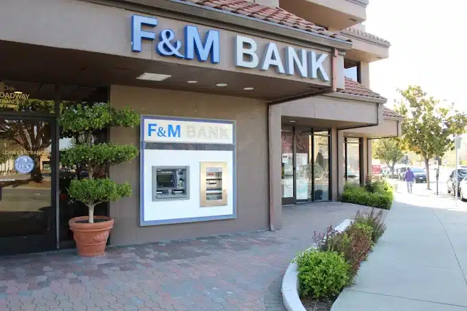 Exterior view of a F&M Bank building with a large sign prominently displaying the bank's name. The entrance features glass doors and a wall-mounted ATM, with potted plants and landscaped greenery along the sidewalk.