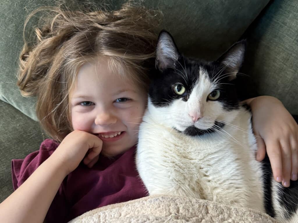 A young child with tousled hair sits beside a black-and-white cat on a green couch, with a beige blanket in the foreground.