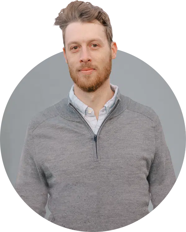 A man with light brown hair and a beard stands against a plain gray background. He is wearing a gray sweater over a collared shirt. The image is framed in a circular shape and is part of the Joybound Team introduction.