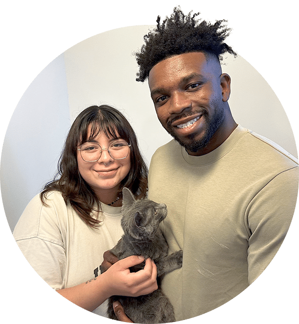 A smiling man and woman in beige shirts hold a gray cat in front of a light wall.