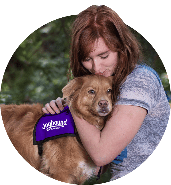 A woman hugs a golden-brown dog wearing a purple Joybound vest, with blurred greenery in the background.