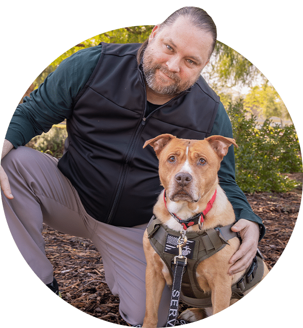 A man kneels beside a tan service dog wearing a harness and leash, with trees and shrubs in the background.