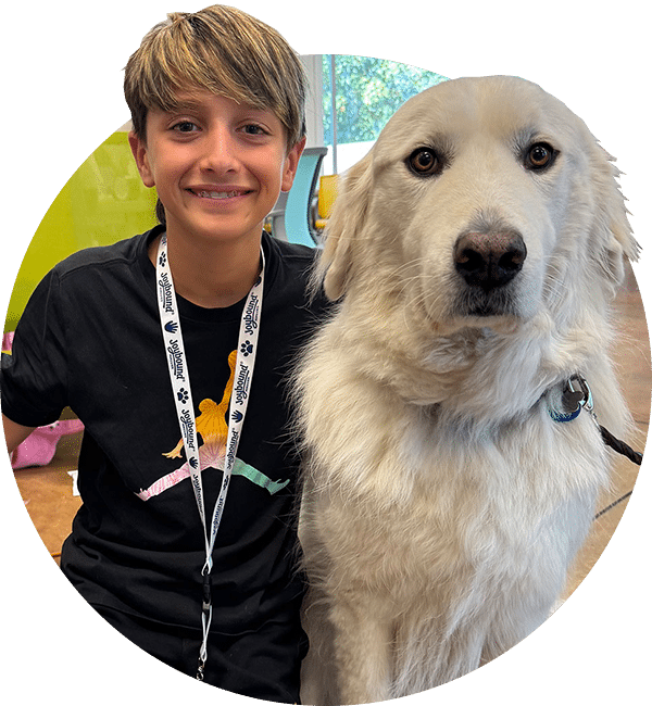 A smiling boy wearing a black T-shirt and lanyard sits beside a large white dog indoors near a window.