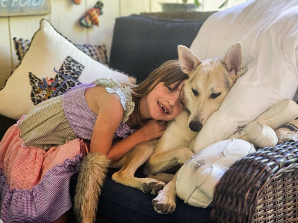 A young girl in a purple dress lies beside a tan dog on a dark sofa, with white pillows and a woven chair in the background.