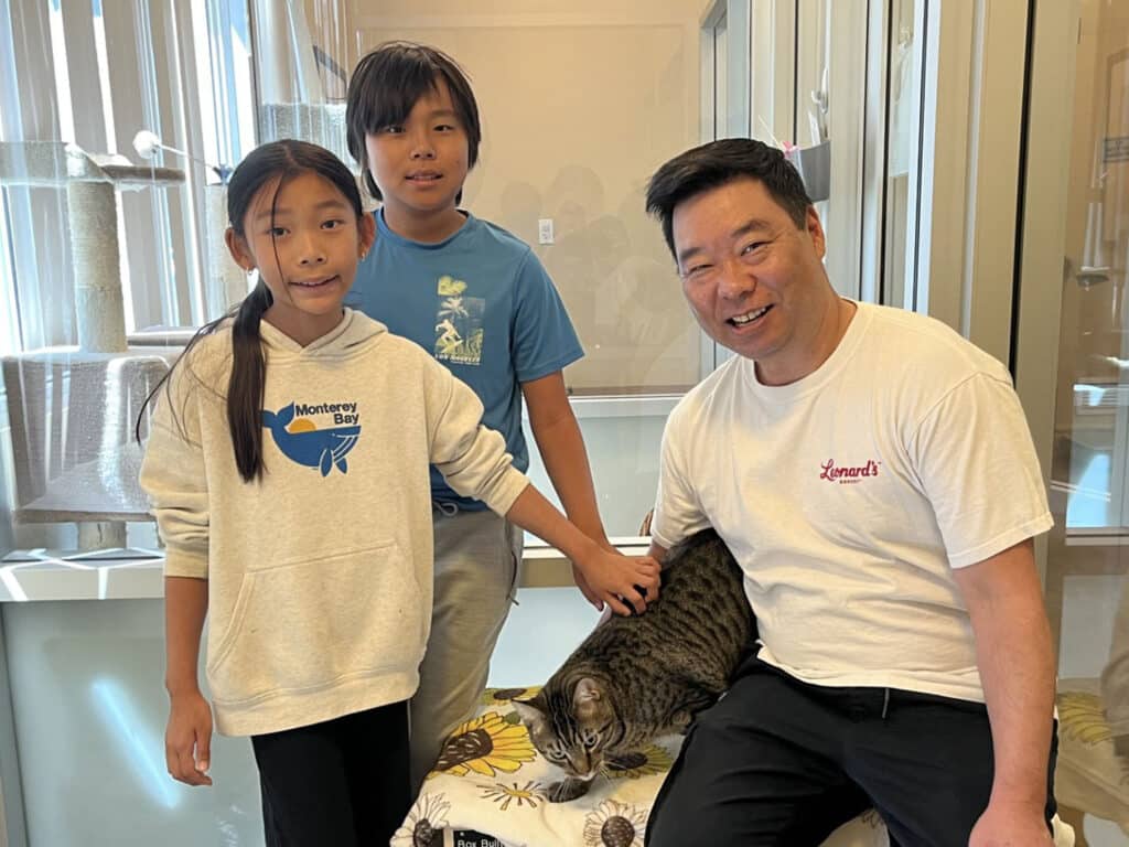 A man in a white T-shirt sits beside two children in a room with glass cases, while a tabby cat rests on a patterned cushion between them.