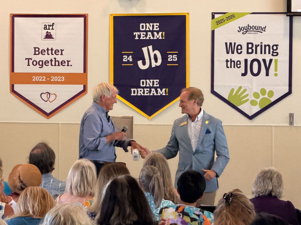 Congressman DeSaulnier shakes hands with a man at the front of a room, with seated attendees and animal welfare banners on the wall behind them.