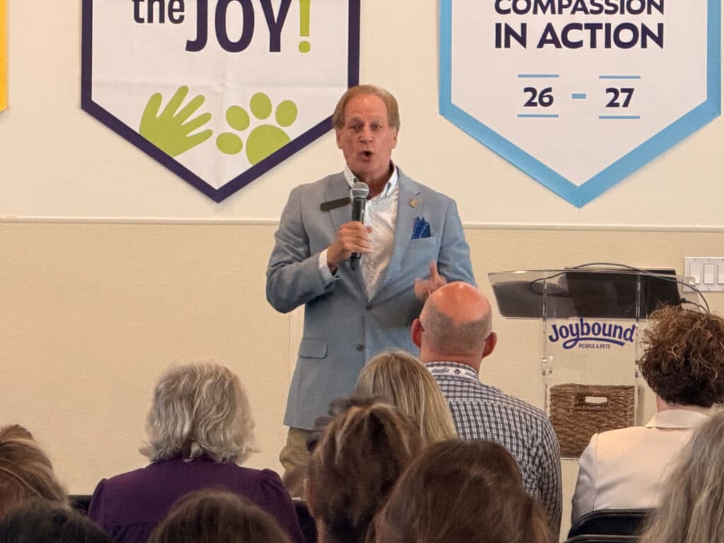 A man in a light blue blazer speaks into a microphone at a podium during a town hall, with seated audience members in the foreground and animal welfare banners on the wall behind him.