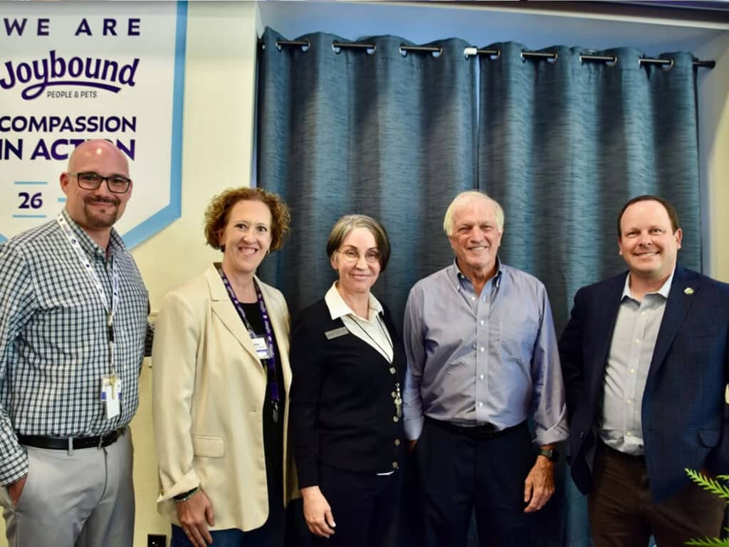 Five adults stand together in front of blue curtains, with a 'We Are Joybound' Compassion in Action banner on the wall at left.