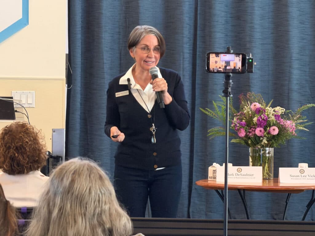 A woman speaking into a microphone stands beside a phone on a tripod and a flower arrangement, with audience members seated in the foreground and dark curtains behind her.