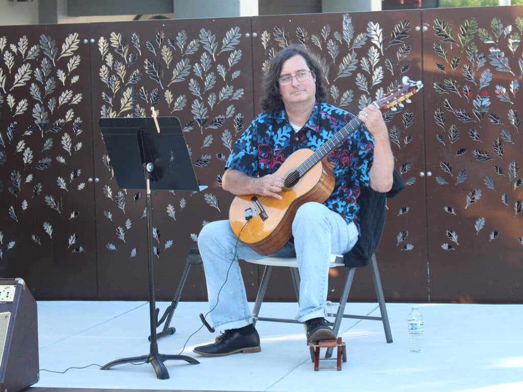 A seated man plays an acoustic guitar on an outdoor stage, with a music stand and decorative leaf-patterned screen behind him.