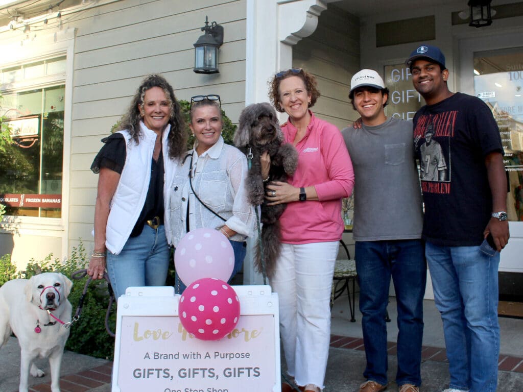 Five people stand outside a storefront holding a dog and a poodle, with a white bulldog at left and a gift shop sign reading "Love for a Cause" in front.