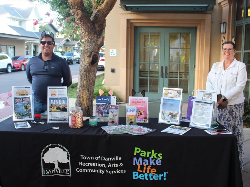 Two people stand behind a Joybound Around Town information table with brochures, flyers, and jars, set outside a building in Danville.