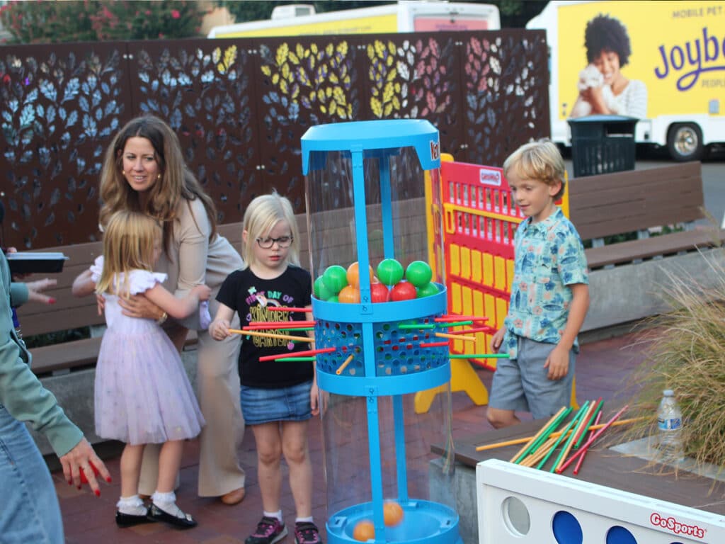 A blue GoSports ring toss game with colorful plastic balls and sticks is set up on a pavement, with a woman and three children standing beside it near a patterned privacy screen.