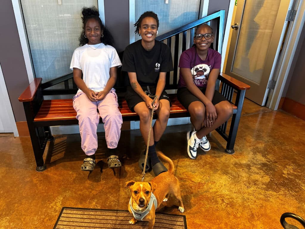 Three girls sit on a bench indoors, with a small brown dog on a leash standing in front of them on a tiled floor.
