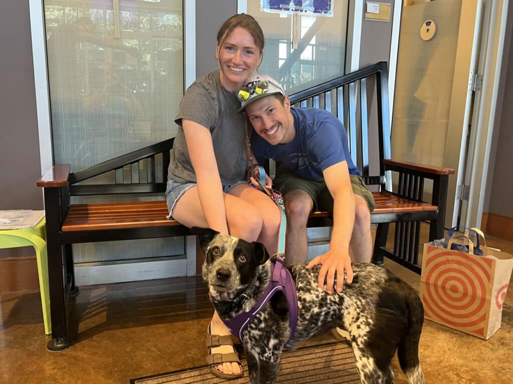A woman and man sit on a wooden bench with a black-and-white dog wearing a purple harness in front of them, inside a lobby with glass walls and a shopping bag nearby.