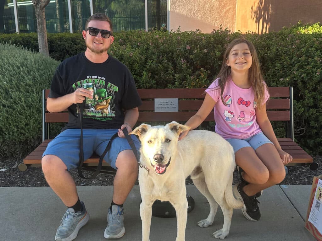 A man and a girl sit on a bench with a white dog standing between them, outdoors in front of shrubs and a building.