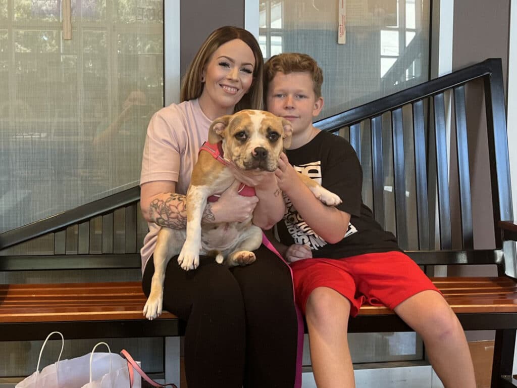 A woman and a boy sit on a bench holding a tan-and-white dog with a pink collar, in front of large windows and dark railings.