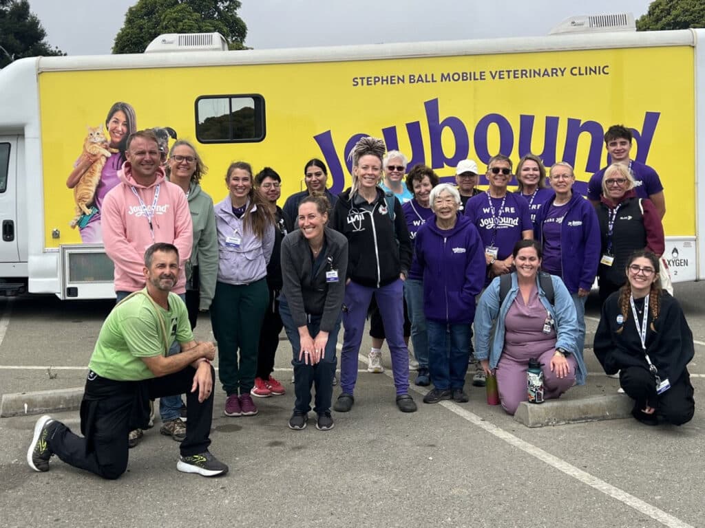 A group of volunteers stands in front of a yellow Stephen Ball Mobile Veterinary Clinic van with large purple lettering, parked on a paved lot.