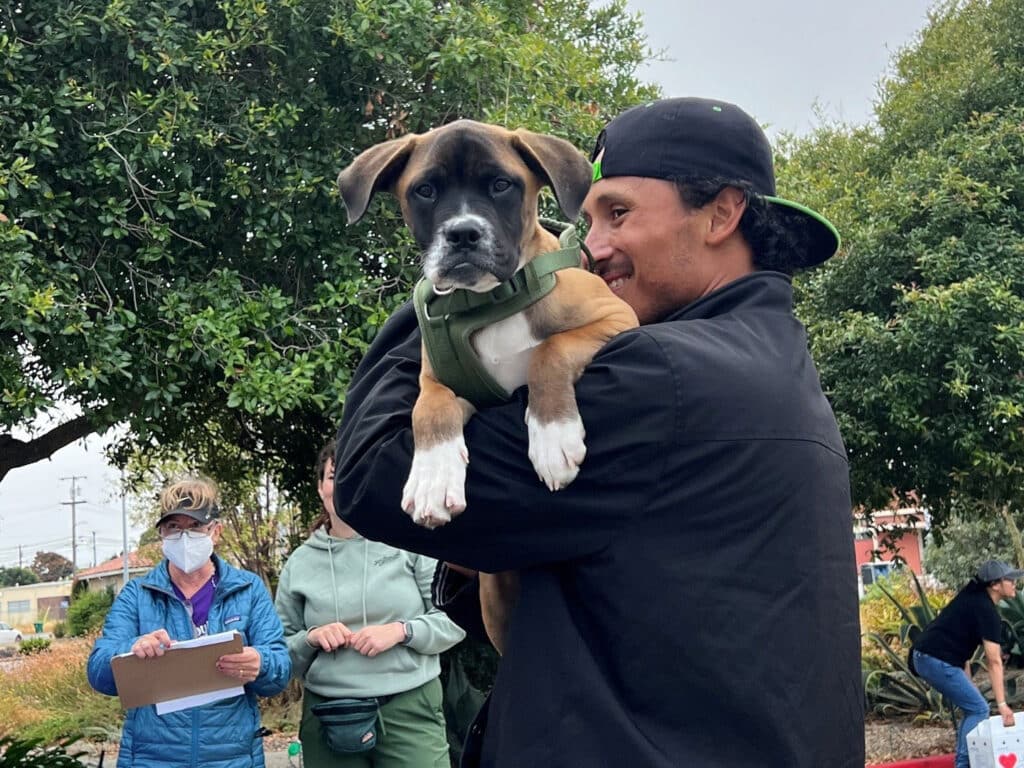A man in a black cap holds a puppy wearing a green harness, with people standing nearby in an outdoor area with trees.