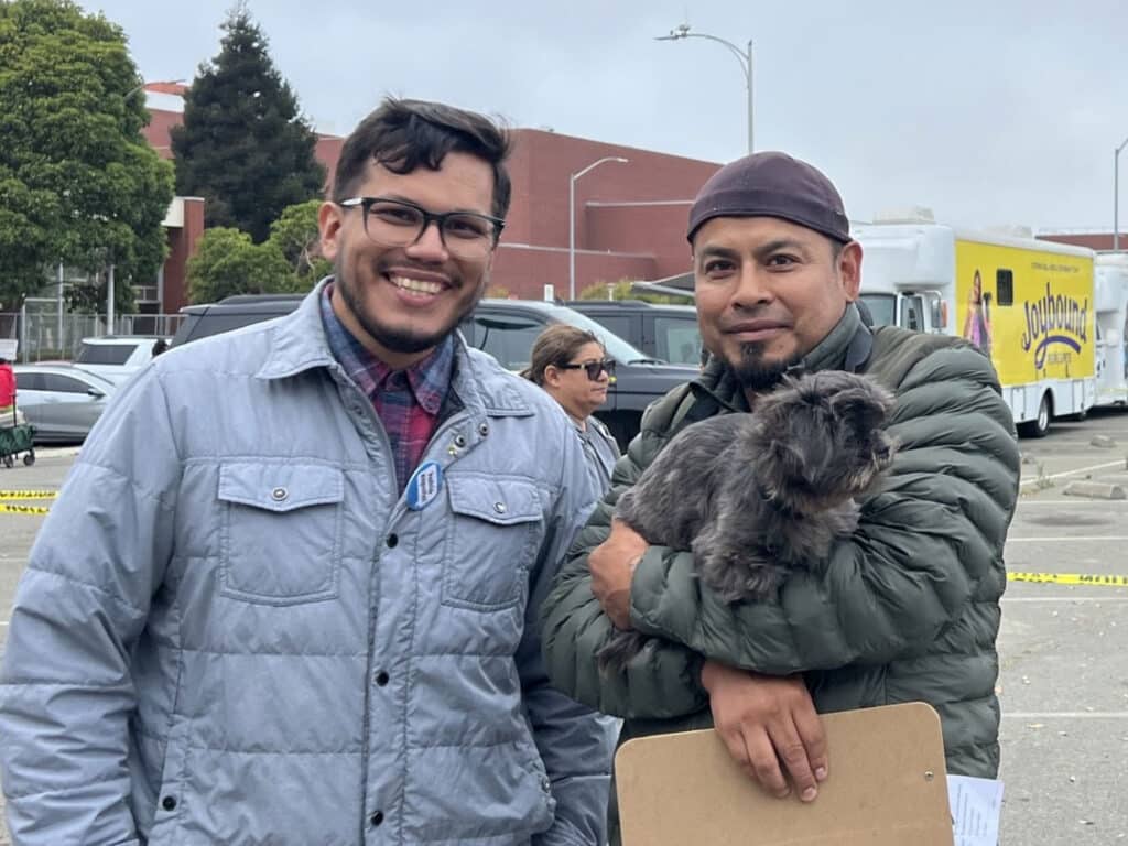 Two men stand outdoors in a parking lot, one holding a small gray dog and a clipboard, with parked cars and a yellow truck in the background.