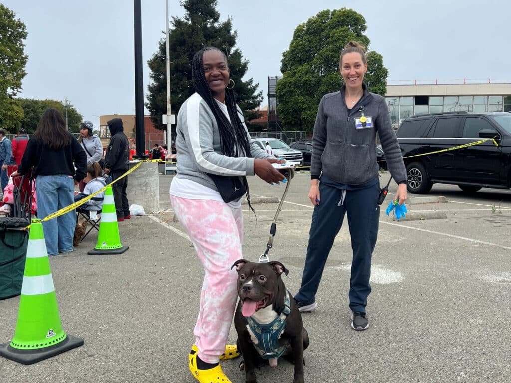 Two women stand with a brown dog on a leash in a parking lot, with green traffic cones, yellow caution tape, and people gathered near parked cars in the background.