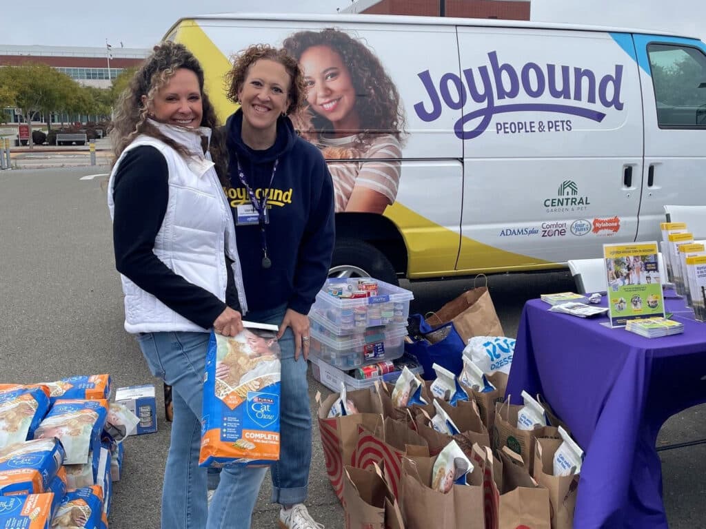 Two women stand beside a Joybound People & Pets van, with bags of pet food, donation supplies, and a purple-covered table in the foreground.